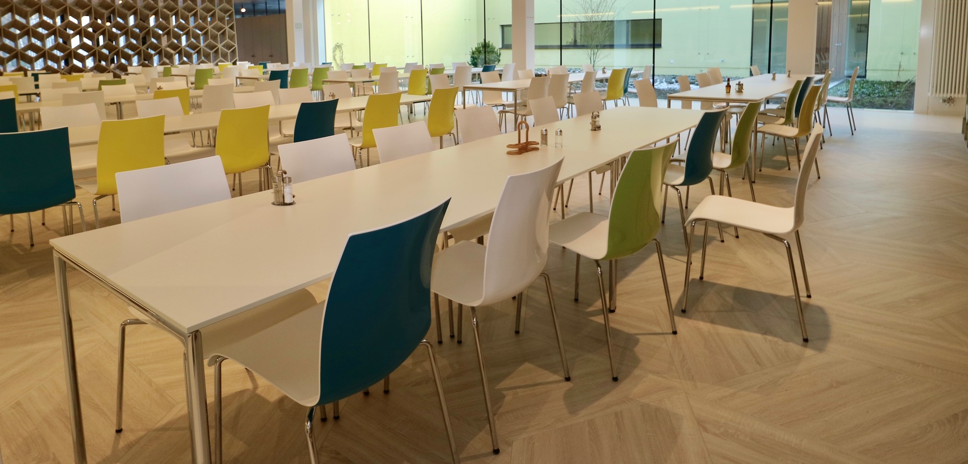 Der Speiseraum des Restaurants. An weißen Tischen sind gelbe und grüne Stühle aufgestellt. Man schaut auf ein großes Fenster zum Innenhof des Raumes